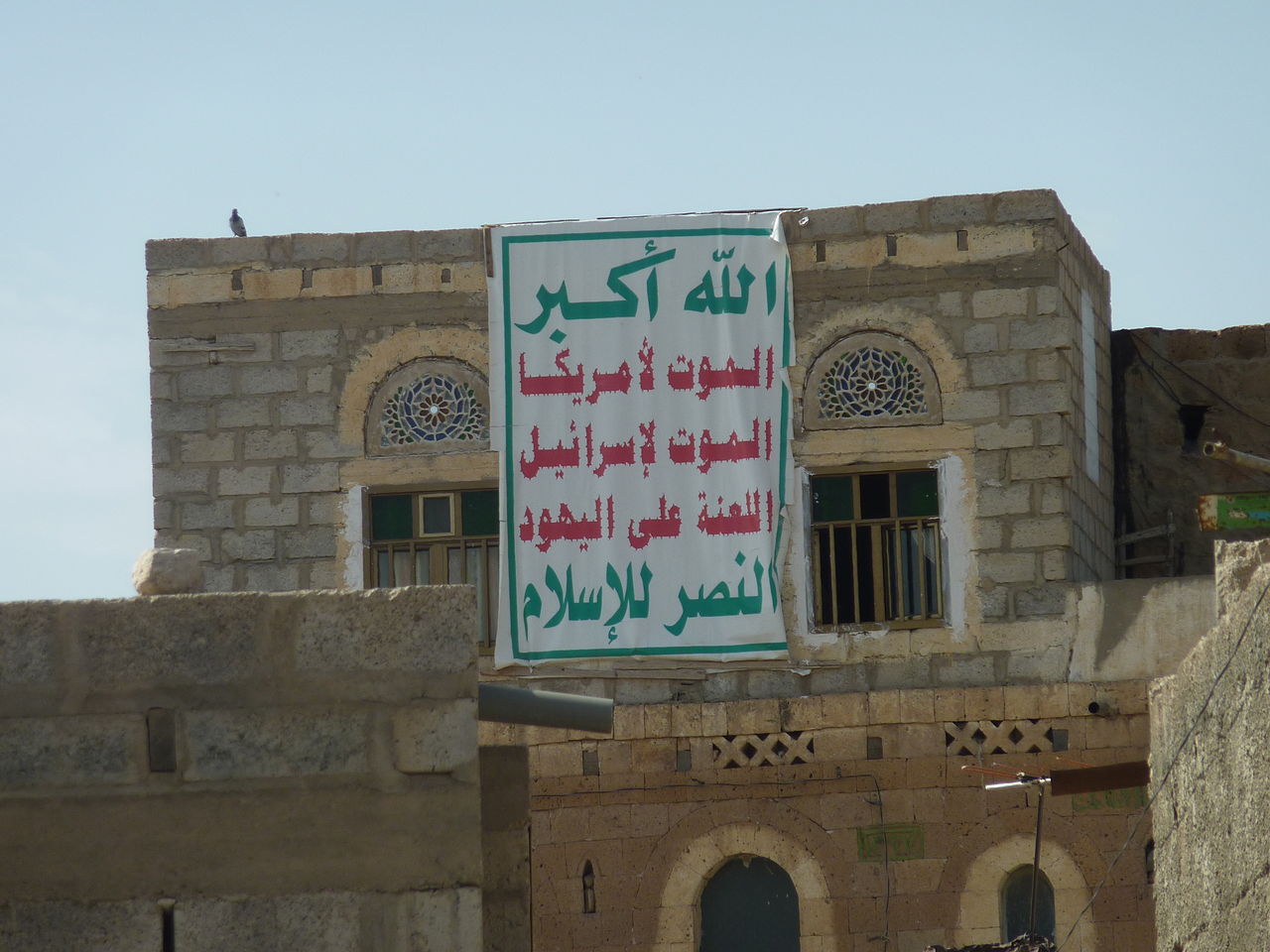 Maison à Dhamar, sud-ouest du Yémen, arborant en septembre 2013 l'emblème des Houthis : « Dieu est grand / Mort à l'Amérique / Mort à Israël / Malheur sur les juifs / Victoire à l'islam ». Crédit : Abdullah Sarhan (licence CC BY-SA 4.0) Maison à Dhamar, sud-ouest du Yémen, arborant en septembre 2013 l'emblème des Houthis : « Dieu est grand / Mort à l'Amérique / Mort à Israël / Malheur sur les juifs / Victoire à l'islam ». Crédit : Abdullah Sarhan (licence CC BY-SA 4.0)